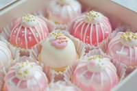 Elegant pink and white cake pops decorated with delicate white drizzle and gold sprinkles arranged in paper cups, showcasing luxurious dessert presentation.