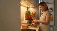 Young woman organizing fresh vegetables and herbs in modern refrigerator with LED lighting for healthy meal preparation and food storage at home.