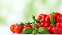 Fresh red bell pepper with cherry tomatoes and basil leaves on white surface with green bokeh background for healthy cooking and nutrition concepts.