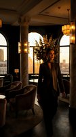 Professional businesswoman in elegant black suit standing in luxury hotel lobby with warm lighting and city skyline view during golden hour sunset.