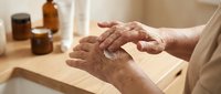 Senior woman applying natural skincare cream to hands at wooden table with amber glass bottles in warm sunlight for anti-aging wellness routine.