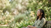 Woman with braided hair sitting on wooden bench in lush garden surrounded by blooming flowers and green foliage, relaxing outdoors in nature.