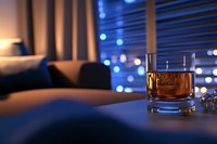 Glass of whiskey on table with blurred city lights through window blinds at night, creating moody atmosphere with warm and cool color contrast.
