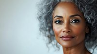 Mature African American woman with gray curly hair and burgundy lipstick smiling at camera against white background. Portrait for beauty, wellness, and lifestyle content.