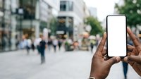 Person with dark skin tone holding smartphone with blank white screen in urban street setting with blurred pedestrians and buildings in background for app mockup or mobile advertising.