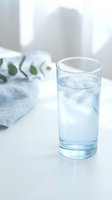 Glass of cold water with ice cubes on white table next to fresh green eucalyptus leaves, bright natural light from window, wellness and hydration concept for health and lifestyle content.