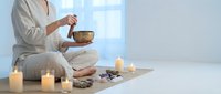 Woman in white clothing practicing meditation with singing bowl surrounded by candles and natural elements in serene wellness space for relaxation.