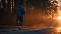 Male runner jogging on forest road during golden hour sunset with warm light filtering through trees creating atmospheric outdoor fitness scene.