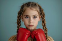 Young female athlete with braided hair wearing red boxing gloves and knit sweater against blue background, portraying strength and determination in youth sports portrait.