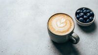 Cappuccino with latte art in gray mug next to bowl of fresh blueberries on concrete surface, ideal for cafe and breakfast content.