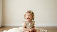 Happy toddler girl with blonde curly hair wearing floral dress, sitting on cream blanket against neutral wall background, smiling at camera for family portraits.