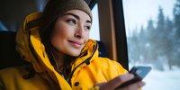 Young woman in yellow winter jacket and knit hat using smartphone indoors with snowy mountain landscape view through window for travel and technology concepts.