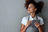 Young African American woman with natural curly hair wearing gray apron and white t-shirt holding digital tablet, looking up thoughtfully against gray background.
