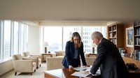 Two business professionals reviewing documents at wooden desk in modern high-rise office with city skyline view, suitable for corporate, finance, and business communication projects.