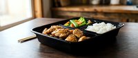 Asian cuisine meal in black compartment tray with fried chicken, steamed rice, and fresh vegetables. Takeout food service.