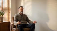 Middle-aged Caucasian man meditating in modern chair by window with wooden blinds creating natural light patterns for mindfulness and wellness content.