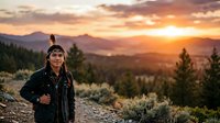 Woman hiker in black jacket stands on mountain trail at golden sunset, overlooking forested valley with distant peaks and warm sky.