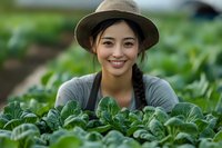 Young Asian woman farmer in hat and apron smiling while tending to organic leafy greens in sustainable greenhouse garden, representing agriculture and farming.