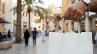 Close-up of male hand holding blank white shopping bag in urban street with palm trees and pedestrians in background for retail and commerce concepts.