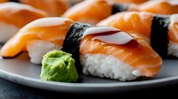 Fresh salmon nigiri sushi with nori strip and wasabi on gray ceramic plate, close up view showing rice texture and glossy fish surface in restaurant setting.