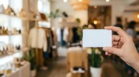 Person holding blank white business card in upscale retail clothing boutique with shelves and merchandise displays blurred in background for branding mockup.