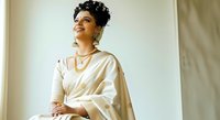 Young woman with dark curly hair and medium skin tone wearing elegant cream silk saree with gold jewelry, seated indoors against neutral background for fashion and lifestyle use.