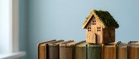 Miniature wooden house with moss roof sitting on stack of old books symbolizing knowledge, education, and sustainable living concepts for real estate.
