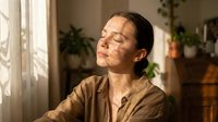 Young Asian woman enjoying warm sunlight streaming through window in cozy home interior with plants creating peaceful morning wellness moment.