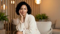 Mature woman with curly black hair and medium brown skin tone, wearing a cream cardigan, resting her chin on her hand while seated on a beige sofa in a modern living room with warm lighting and potted plants.