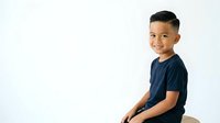 Young boy, approximately 6-8 years old, Southeast Asian ethnicity, wearing dark navy blue t-shirt, sitting against white background, smiling at camera with confident expression, studio portrait style.