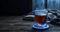 Steaming hot tea in glass mug on wooden table with cozy blanket and book creating warm atmospheric scene for relaxation and comfort at home.