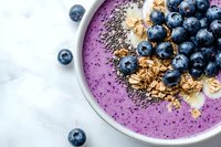 Purple smoothie bowl topped with fresh blueberries, granola, banana slices and chia seeds on marble background, healthy breakfast concept for food blogs.