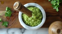 Fresh homemade pesto sauce in stone mortar with wooden pestle on rustic wooden background with herbs and ingredients.