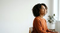 Young woman with dark curly hair and medium skin tone sitting indoors, wearing rust-colored sweater, looking upward thoughtfully near window with plant.