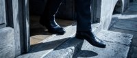 Man in formal black shoes and dark trousers standing on sunlit concrete ground between pillars, casting sharp shadows. Suitable for business, professional, and corporate concepts.