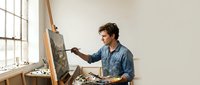Young male artist painting on canvas easel in bright studio workspace with natural light from large windows for creative projects and artistic expression.