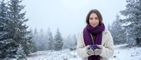 Young Caucasian woman in winter coat and purple scarf holding snow in snowy forest, smiling warmly at camera on cold foggy day.