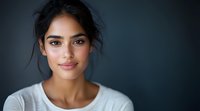 Young mixed race woman with natural makeup and messy bun hairstyle wearing white casual top smiling confidently against dark background in studio portrait.