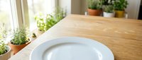 Empty white plate on wooden table with potted plants and natural window light, suitable for food styling, restaurant menus, and culinary content.