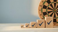 Wooden blocks with checkmarks arranged in ascending order next to dartboard, representing progress, achievement, and success in business goals and performance metrics.