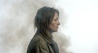 Middle-aged Caucasian woman with disheveled dark hair in profile view against smoky gray background, conveying contemplation and introspection.