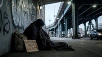 Homeless man sitting under urban bridge wrapped in blanket, holding cardboard sign, backpack nearby, gritty city underpass with graffiti.