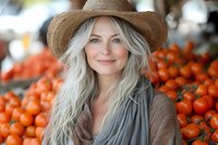 Mature woman with long silver hair wearing straw hat smiling at farmers market with fresh oranges in background, natural lifestyle portrait for wellness and aging.