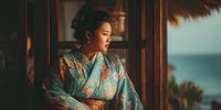 Young Asian woman in traditional floral kimono gazing thoughtfully through wooden window frame with ocean view in warm golden light for cultural lifestyle content.