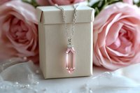 Elegant pink crystal pendant necklace on silver chain with gift box against soft rose background, showcasing delicate jewelry piece for special occasions.
