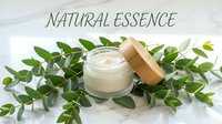 Natural essence skincare cream in glass jar with wooden lid surrounded by fresh green leaves on white surface.