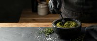 Matcha green tea powder in dark bowl with bamboo whisk on slate surface, traditional Japanese tea ceremony preparation for culinary and wellness use.