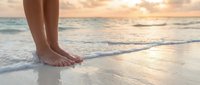 Woman's bare feet standing on wet sand at ocean shore during golden hour sunset with gentle waves washing over beach creating peaceful vacation moment.