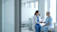 Female doctor in white coat consulting with elderly Caucasian woman patient in hospital room. Healthcare professional providing medical care and support.