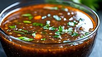 Rich brown sauce with herbs and vegetables in glass bowl, savory condiment for cooking and food preparation.